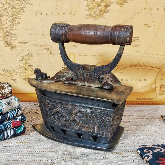 A vintage charcoal iron with a dark brown wooden handle, displayed on a woven textile surface with a vintage map in the background.