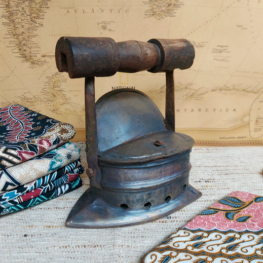 A vintage dark metal charcoal iron with a curved soleplate and a cylindrical handle, displayed on a textured fabric surface with patterned textiles and a vintage map backdrop.