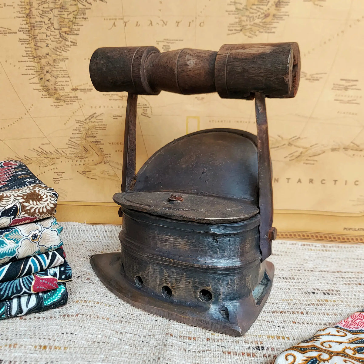 A vintage dark metal charcoal iron with a wooden handle, displayed on a woven textile surface in front of a map backdrop. Folded patterned fabrics are partially visible on the left side.