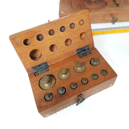 A wooden box with opened lid, containing ten small brass weights inside, shown on a white background with a yellow measuring tape visible in the background.