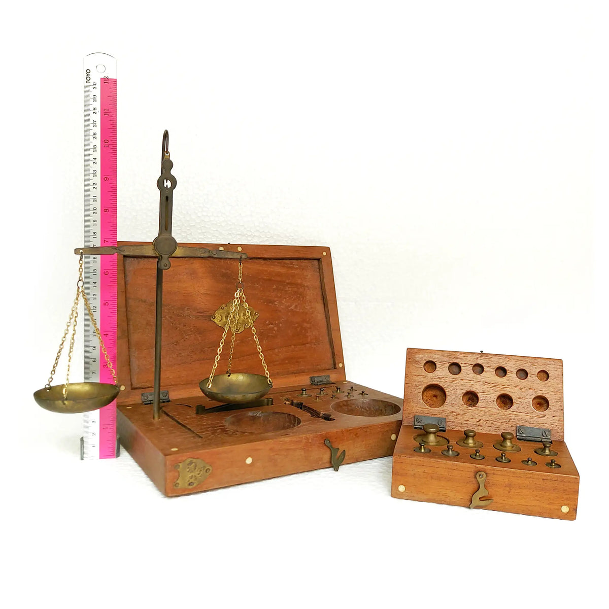 A vintage brass balance scale set with a smaller box containing weighing scales on the right. A pink-and-white ruler is standing behind the left unit for size reference, against a plain white background.