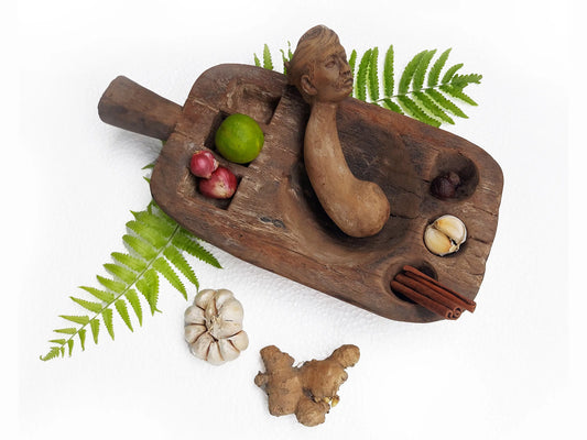 A rustic carved wooden mortar and pestle with a sculpted human head, styled with green fern leaves and natural ingredients including garlic, ginger, cinnamon sticks, a lime, and small red onions on a white background.
