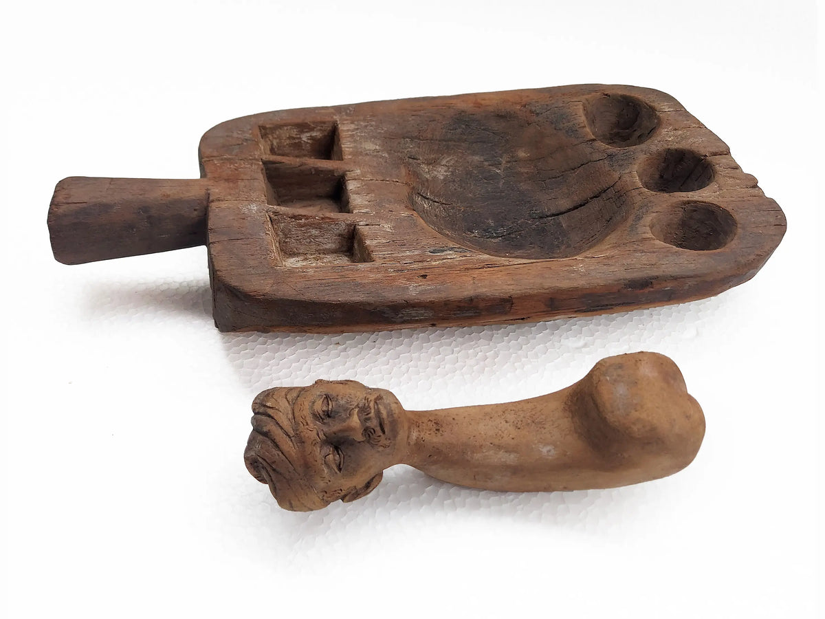 A carved wooden mortar with circular and square compartments, and pestle with a sculpted human figure head, photographed on a plain white background.