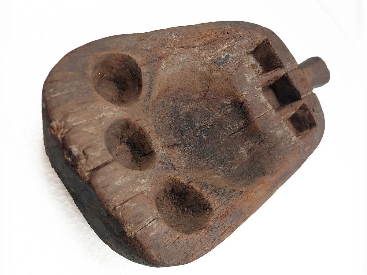 A hand-carved wooden pestle with four circular and square indentations and a protruding peg, shown on a plain white background.