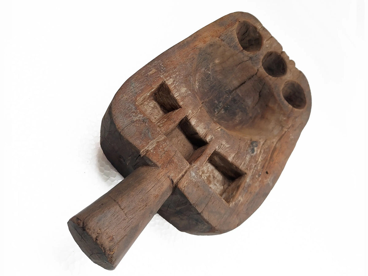 A rustic, hand-carved wooden pestle with square and circular compartments, shown on a plain white background.
