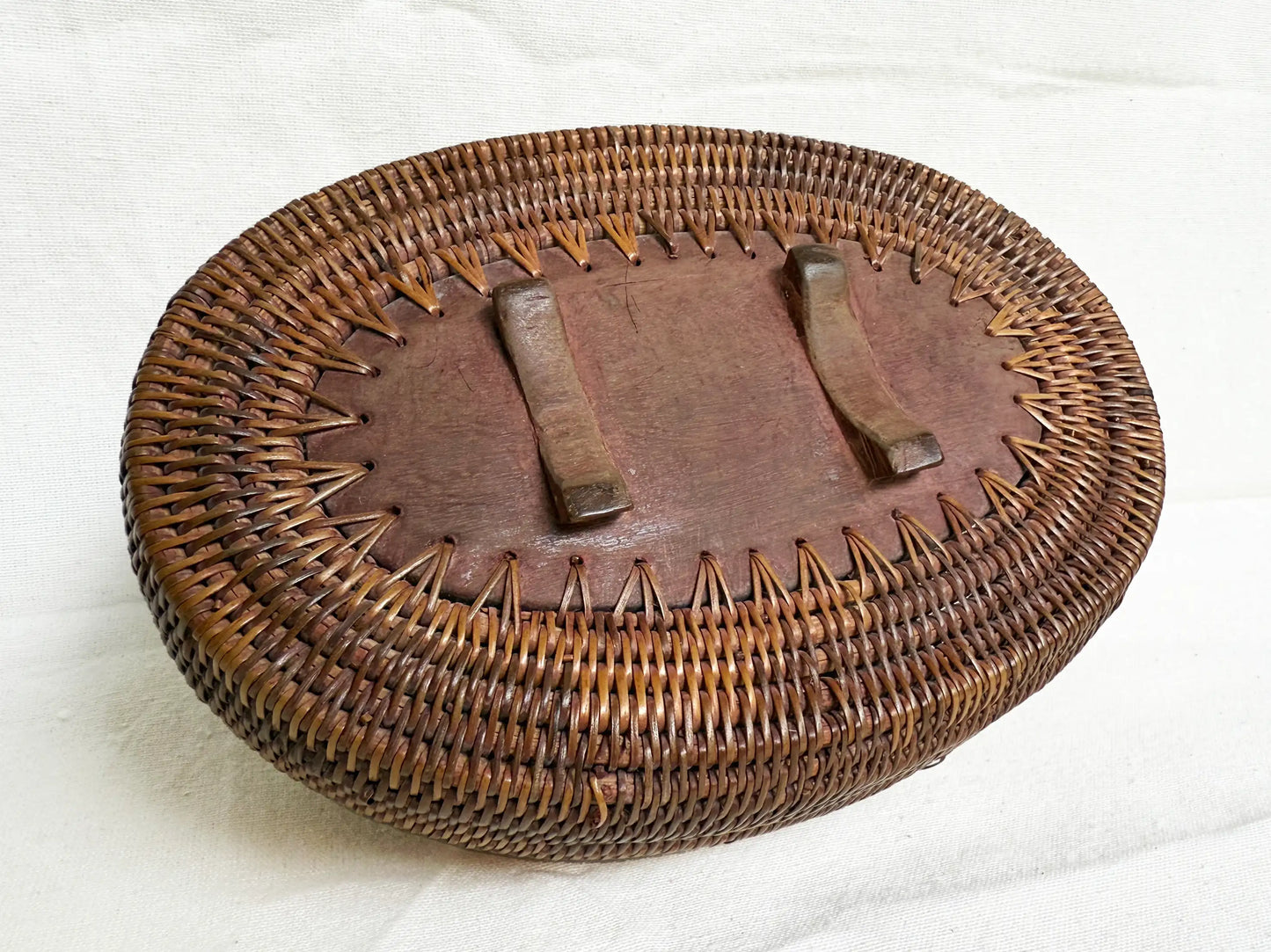 An oval woven rattan storage basket bottom part with a wooden base featuring two short legs, photographed on a white fabric background.