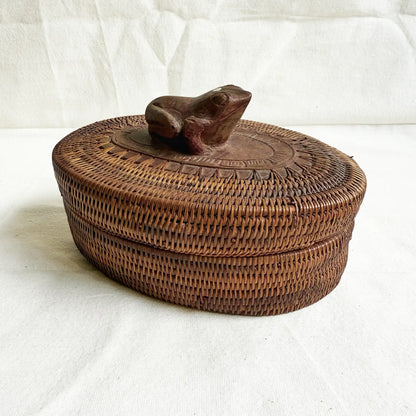 An oval woven basket with a lid, featuring a carved frog-shaped handle on top, set against a white fabric backdrop.