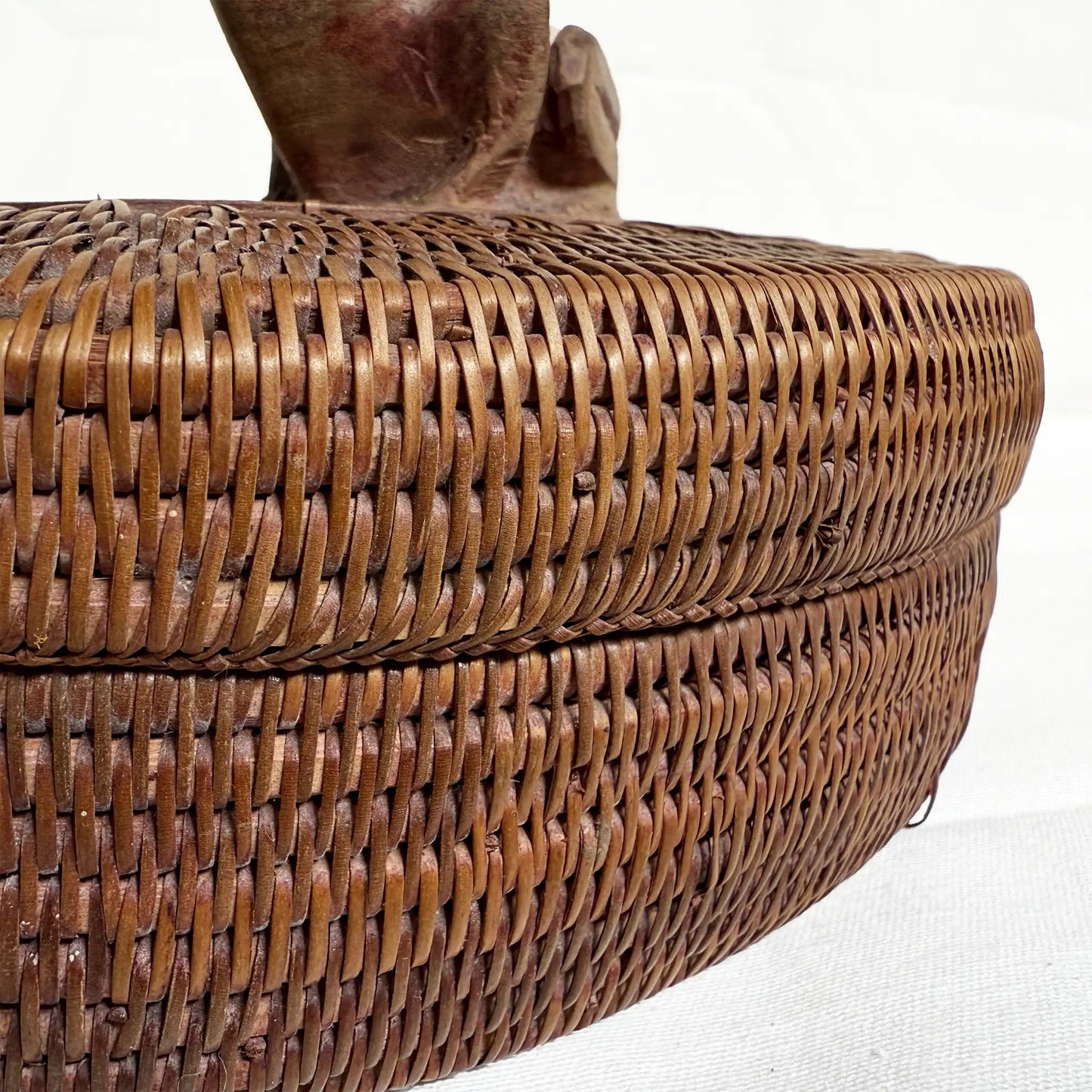 Close-up of a brown woven basket with a lid, showing the detailed interlaced texture and a portion of the handle against a white background.