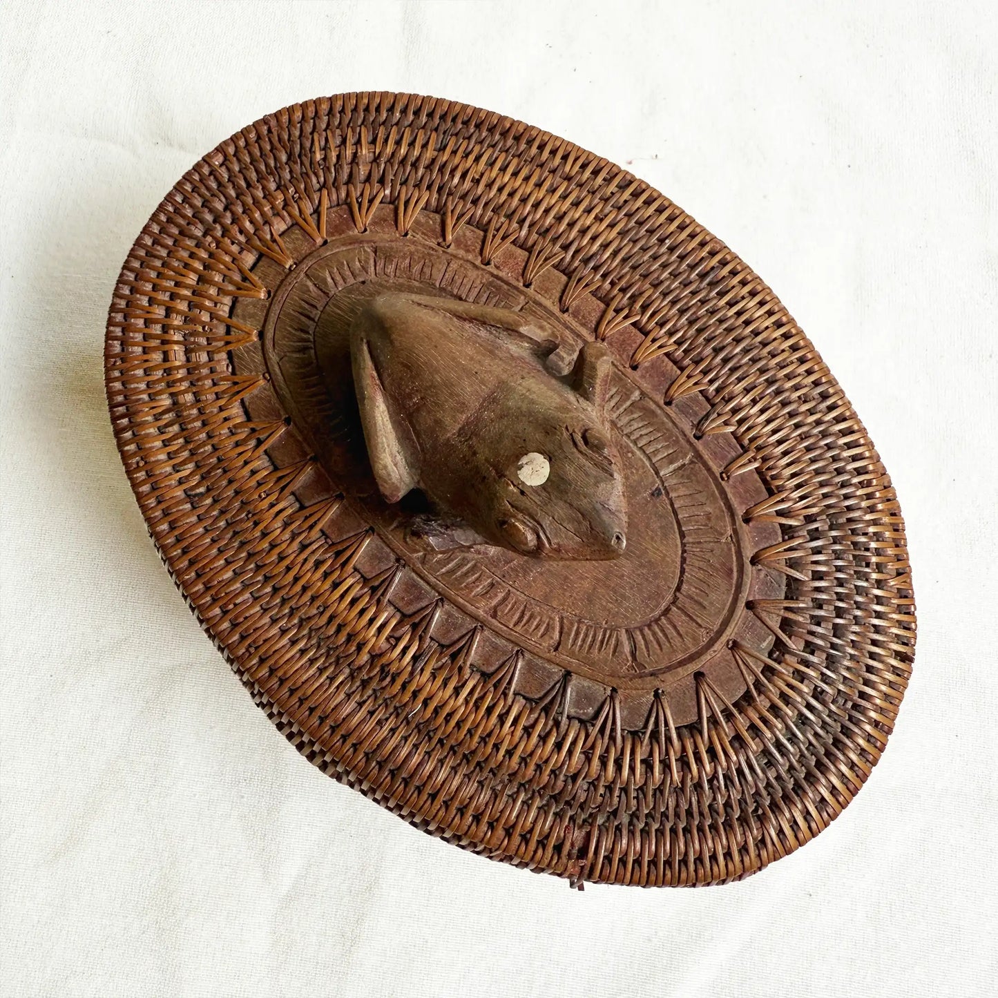 A round, hand-woven brown basket with a carved wooden frog mounted in the center, photographed from above on a light fabric background.