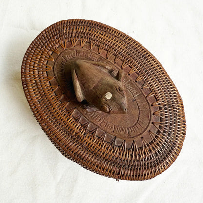 A round, hand-woven brown basket with a carved wooden frog mounted in the center, photographed from above on a light fabric background.