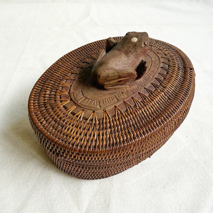 A brown woven basket with a lid, featuring a carved wooden frog figure on top and a circular carved wooden base around the frog, photographed on a white fabric background.
