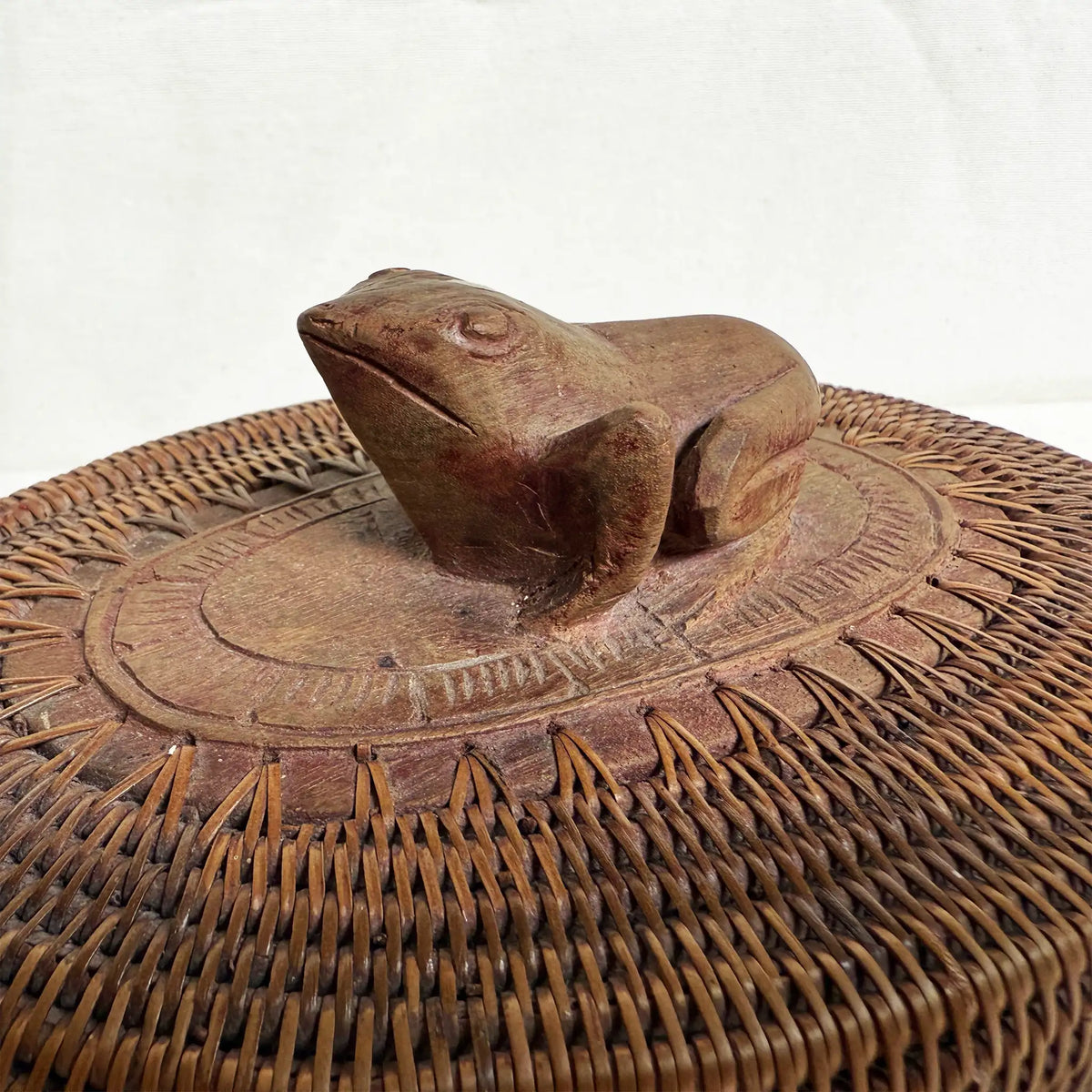 A close-up of a woven basket featuring a carved frog-shaped handle on top, shown against a plain light background.