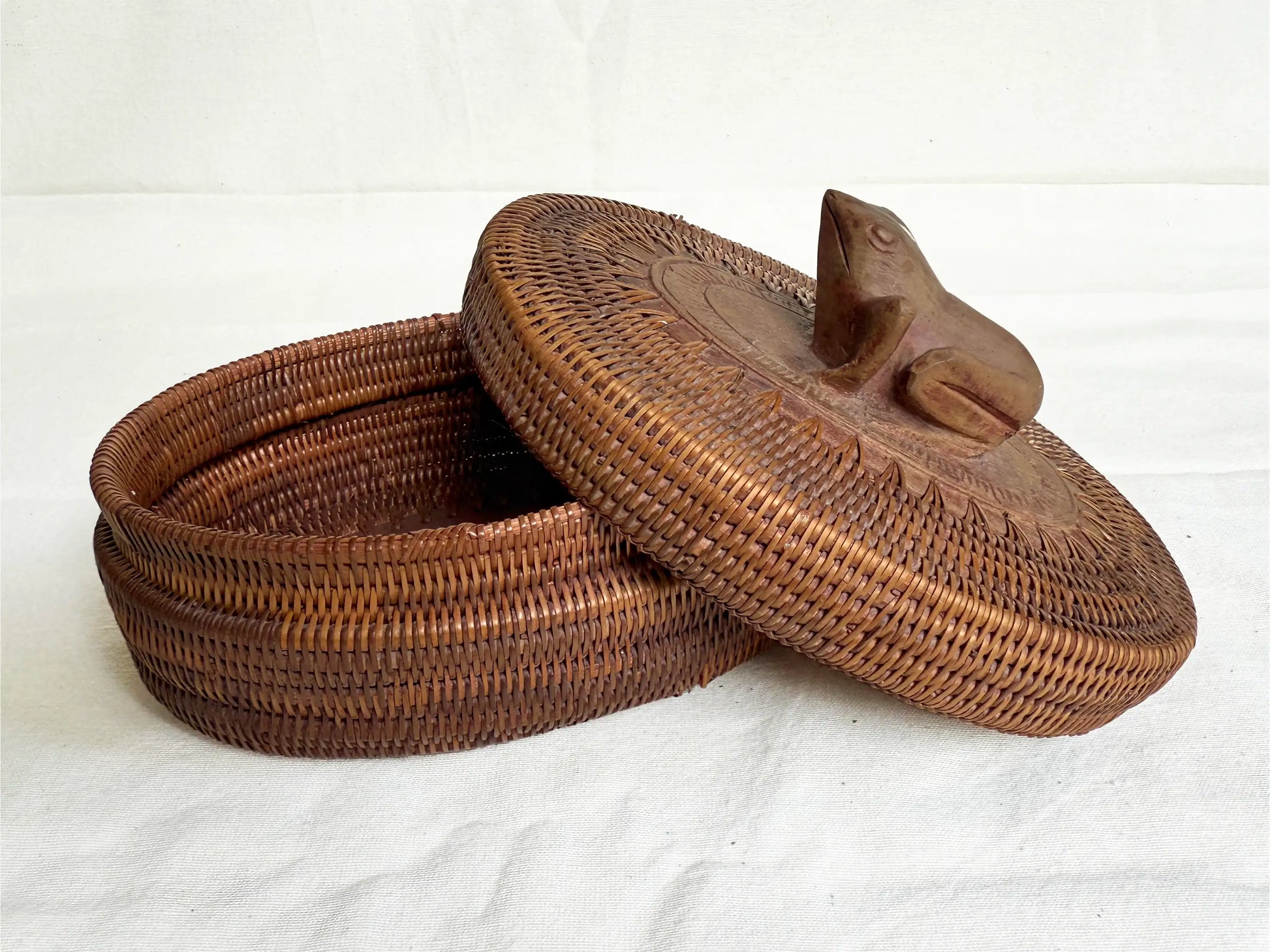 A brown woven basket with a matching lid featuring a carved wooden frog topper, set against a white fabric background.