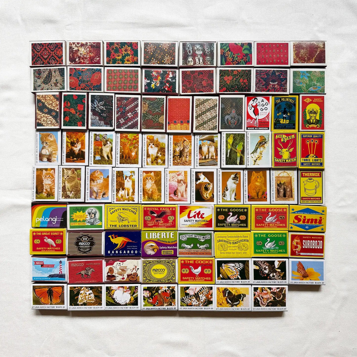 Vintage matchbook covers arranged in a grid on a white fabric background. The matchbooks feature many different designs, colors, and printed illustrations, including animals, landscapes, and logos 