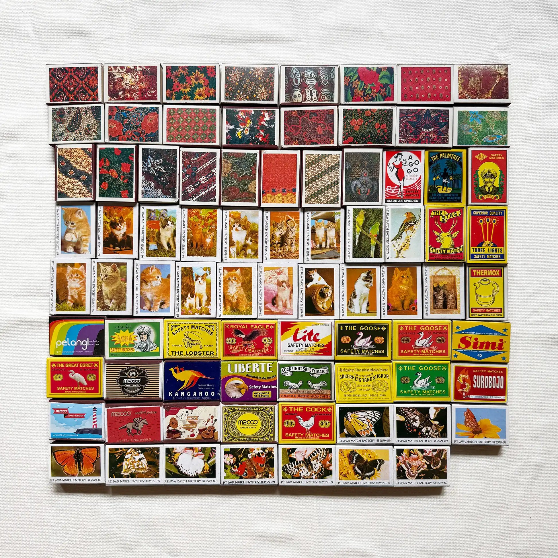 Vintage matchbook covers arranged in a grid on a white fabric background. The matchbooks feature many different designs, colors, and printed illustrations, including animals, landscapes, and logos 