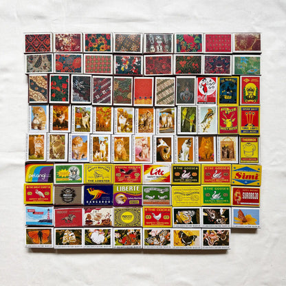 Vintage matchbook covers arranged in a grid on a white fabric background. The matchbooks feature many different designs, colors, and printed illustrations, including animals, landscapes, and logos 
