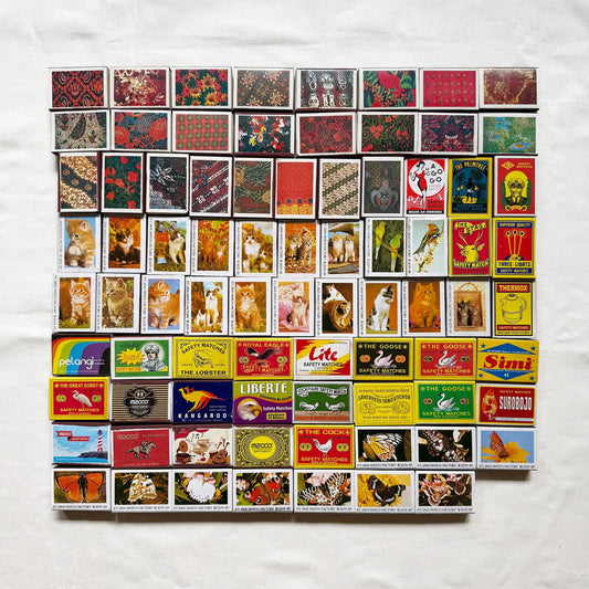 Vintage matchbook covers arranged in a grid on a white fabric background. The matchbooks feature many different designs, colors, and printed illustrations, including animals, landscapes, and logos 