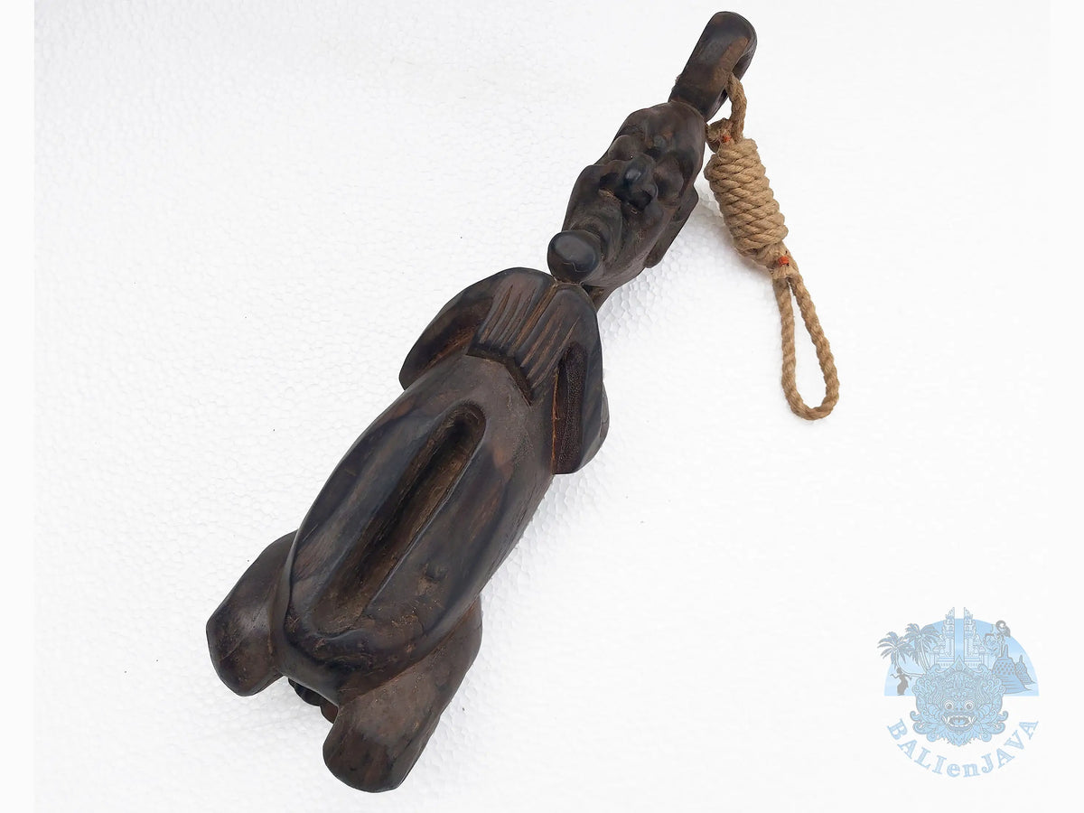 A dark carved wooden slit drum with a human shape and rope loop, photographed on a plain white background.