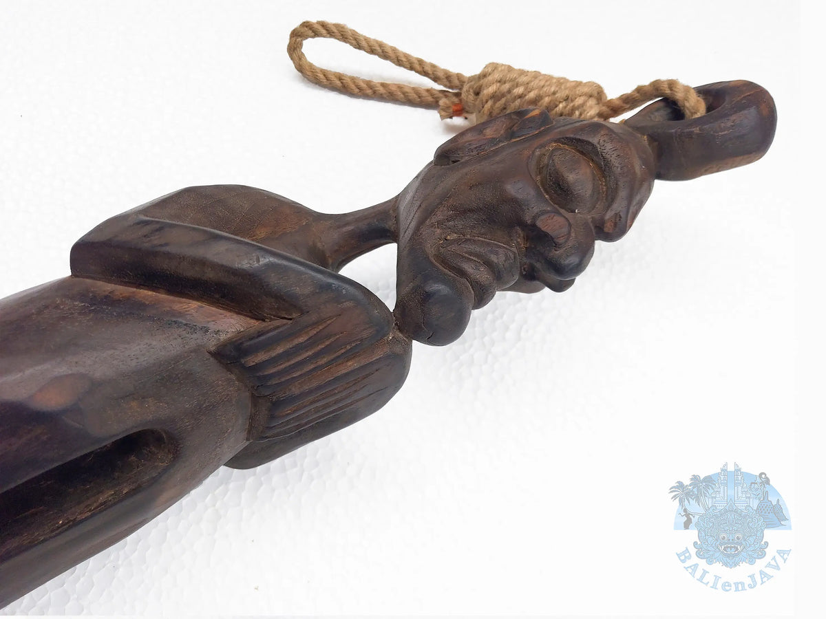 A close-up of a dark carved wooden object with a rope loop at the top, photographed against a plain white background. 