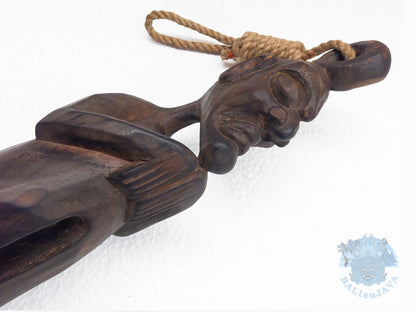 A close-up of a dark carved wooden object with a rope loop at the top, photographed against a plain white background. 