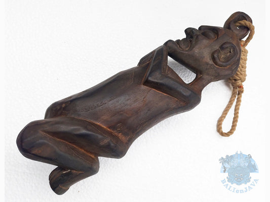 A dark brown carved wooden slit drum of a reclining figure with a cutout face, featuring a natural fiber hanging cord, displayed against a white background.