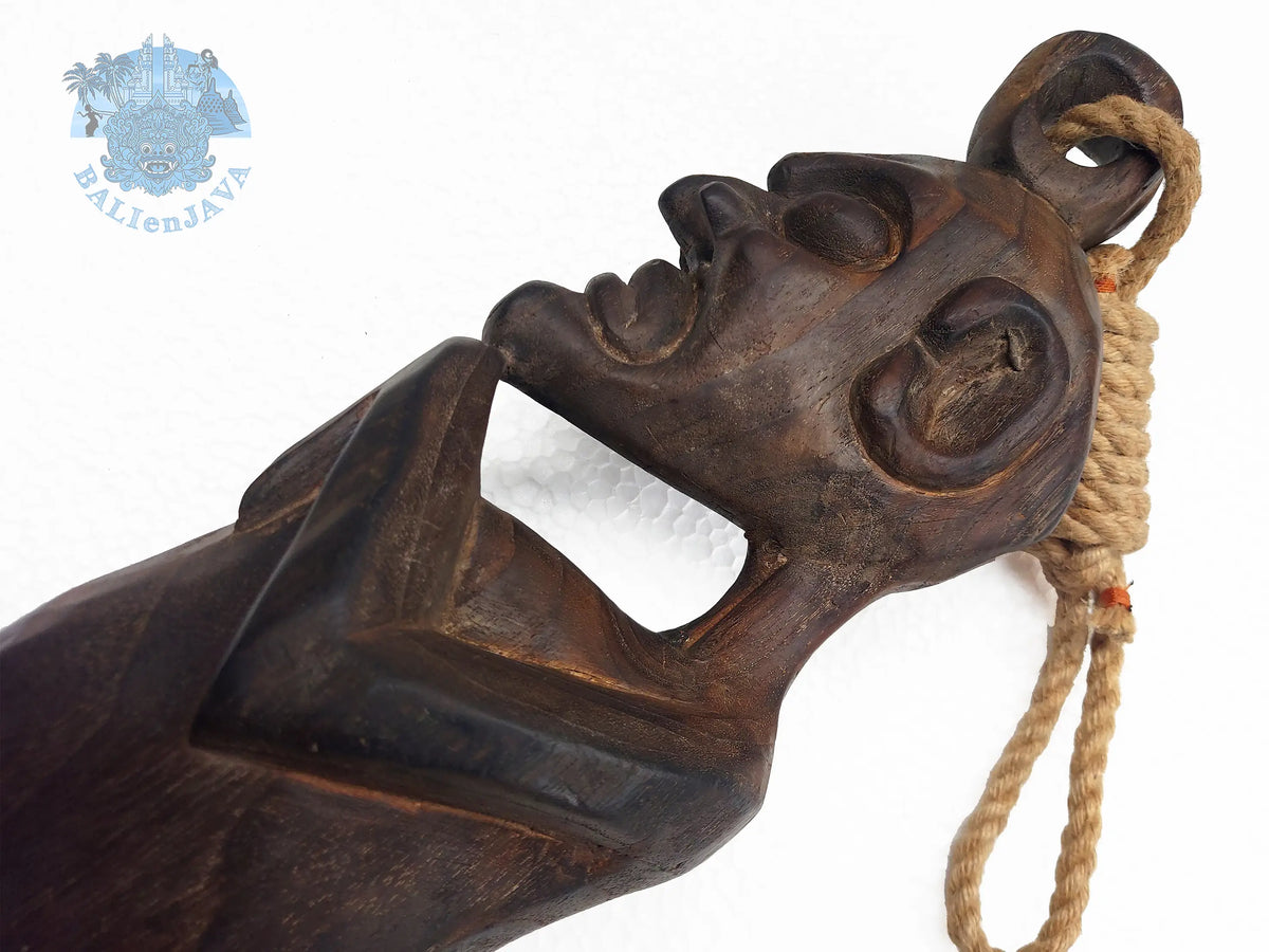 A close-up of a dark carved wooden  slit drum instrument, featuring a stylized face and a beige rope loop, shown against a plain white background.